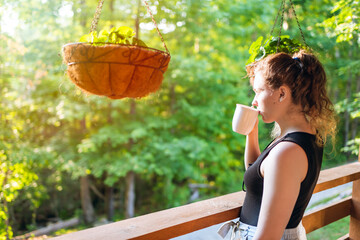 Hanging potted plants in summer with woman standing on porch of house in morning wooden cabin cottage drinking tea or coffee