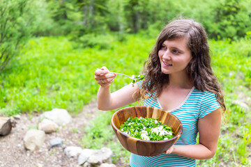 Happy young woman eating kale salad from large wooden bowl with bokeh background of green Utah...