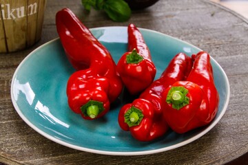 Close up of group of isolated long red bell peppers on blue china dish and round wood table. Blurred basil pot background.