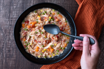 Woman’s hand with black metal spoon, Puerto Rican Chicken Rice Soup in a black stoneware bowl on a dark wood table, orange cloth napkin