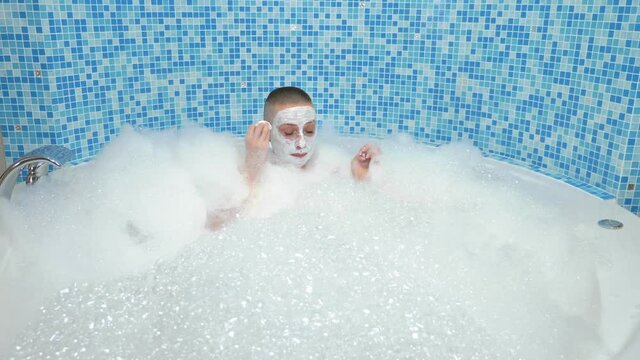 a bald young woman with a mask on her face lies in a bath with foam, she smiles and blows on the foam
