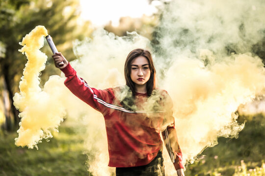 Young asian woman holding yellow colorful smoke bomb on the outdoor park. Yellow smoke spreading