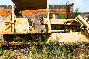 Photograph of an old abandoned yellow tractor, in the middle of a meadow with high brushwood. In the background a wall and the blue sky.