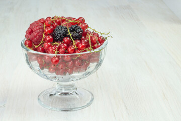 Composition of berries (red currants, blackberries, raspberries), fruit in a glass bowl on a white table.