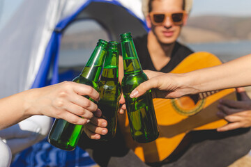 Close up of young man with guitar in front of tent at the beach holding beer and cheering with his...