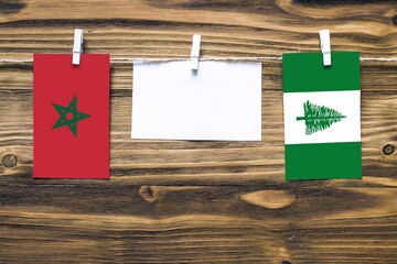 Hanging flags of Morocco and Norfolk Island attached to rope with clothes pins with copy space on white note paper on wooden background.Diplomatic relations between countries.