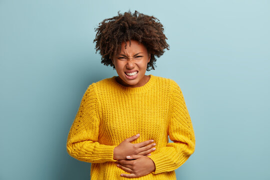 Displeased dark skinned woman suffers from stomachache, period cramps, keeps hands on belly, wears yellow knitted sweater, smirks face, stands against blue background. People and sickness concept