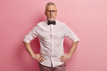 Half length shot of self confident mature male entrepreneur keeps both hands on waist, looks seriously at camera, ready for work, wears formal clothing, optical glasses for vision correction