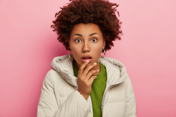 Candid shot of surprised woman in winter outfit puts hand on dropped jaw, stares with astomishment and disbelieving gets shocking news isolated over pink background. People, emotions, face expressions