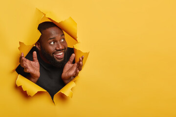 Glad black man spreads palms sideways, looks happily aside, notices something pleasant, has thick bristle, white teeth, poses in torn hole of yellow background, makes choice looks entertained, excited