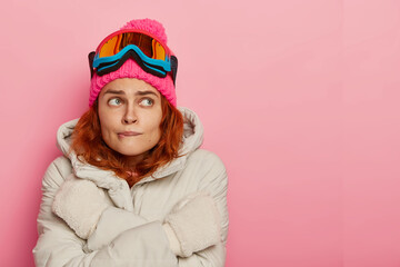 Girl skier feels cold, trembles and bites lips, wears warm winter outerwear, has active recreation during winter time, tries to warm, stands against pink studio wall, copy space for your advertising