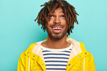 Headshot of happy man with dreads, has positive expression, focused above, wears sailor jumper and yellow raincoat, isolated over blue background, enjoys rainy autumn weather. Cheerful teenager