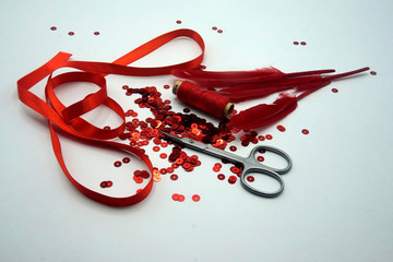 Red sequins, spool of red thread, needle, scissors, red ribbon and red feathers on white background.