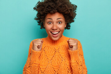 Charismatic cute African American lady cheers victory, raises clenched fists, celebrates something, smiles broadly, shows white teeth, wears knitted orange sweater, being extremly excited feels upbeat