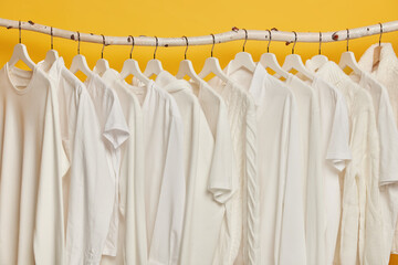 Same white clothes on wooden racks in closet. Collection of clothing on hangers, isolated over yellow background. Dressing, style and fashion concept. Selection of solid apparel. Home wardrobe
