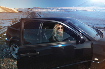 Fashion style wide angle young stylish attractive girl with bright red lips in a leather jacket and sunglasses sits in a retro 90s car with an open door on nature in the mountains at sunset