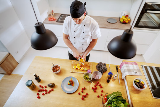 Top View Of Dedicated Creative Chef In Uniform Cutting Orange To Decorate Salmon. On Kitchen Counter Are Vegetables, Spices And Plate With Salmon.