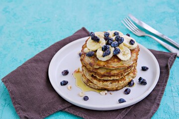 Banana oat pancakes with hazelnuts, sliced banana, honey and honeysuckle on a white plate on a turquoise concrete background. American food. Baking with oatmeal.