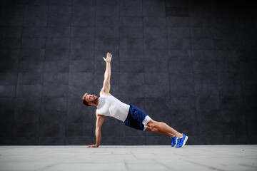 Handsome sporty caucasian man in shorts and t-shirt stretching his arm in plank position in front of gray wall.