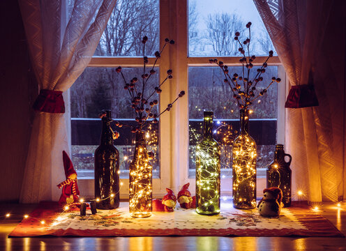 Cute and warm Christmas decoration set with vintage beer bottles and wine bottles filled with micro led party lights, behind the window is countryside forest. Bottles vases for red artificial berry.