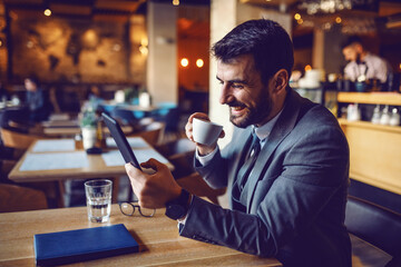 Handsome smiling caucasian bearded businessman in suit sitting in cafe, drinking coffee and using tablet. - Powered by Adobe