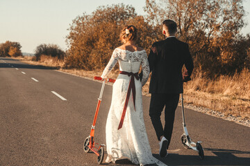 Wedding couple riding a on scooters along the road outside the city at sunset. Shoot from the back....