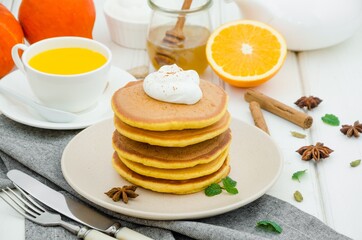 Stack of spicy pumpkin pancakes on a plate with honey and whipped cream on a white wooden background.  Rustic style. Fragrant autumn breakfast. Breakfast for Halloween.