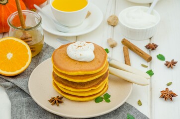 Stack of spicy pumpkin pancakes on a plate with honey and whipped cream on a white wooden background.  Rustic style. Fragrant autumn breakfast. Breakfast for Halloween.