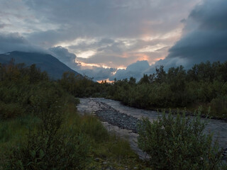 wild river with birch tree forest and mountains in sunset light. Lapland nature landscape in summer, moody sky, pink clouds.