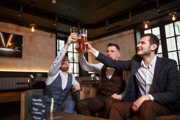 A cheerful company of young stylish guys in suits of three people clink glasses with beer at a party in a pub.