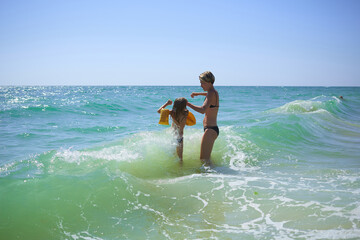 Summer happy family of six years blonde child playing and jumping water waves embracing woman mother in sea shore beach