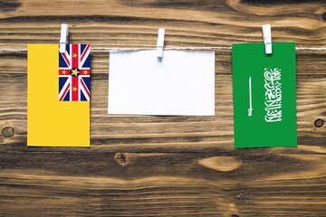 Hanging flags of Niue and Saudi Arabia attached to rope with clothes pins with copy space on white note paper on wooden background.Diplomatic relations between countries.