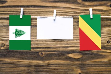 Hanging flags of Norfolk Island and Republic Of The Congo attached to rope with clothes pins with copy space on white note paper on wooden background.Diplomatic relations between countries.