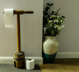 Upcycled washing dolly re-purposed as a toilet roll holder in bathroom setting. with loo rolls. Floral display in vase to one side with pastel wall background and wooden flooring. - 300006221