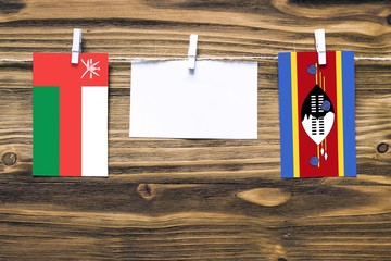 Hanging flags of Oman and Swaziland attached to rope with clothes pins with copy space on white note paper on wooden background.Diplomatic relations between countries.