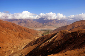 Panoramic view of the Tibetan Plateau showing brown mountains against a blue sky covered by white...