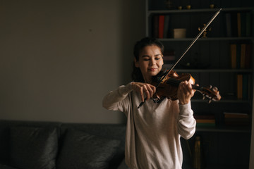 Lovely young woman plays the violin at home, in a room with a modern cozy interior. Girl is...