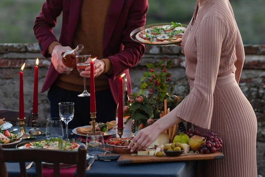 Young couple is waiting for guest to come to beautifully decorated dinner party. Candles and fresh fruits as decor, delicious snacks, pizza, wine. Relaxed atmosphere, festive mood