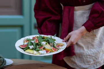 Young slim girl in red fress is holding white plate with delicious and healthy salad: green arugula, figs, pears, blue cheese. Tasty and low calories lunch. Bright colors, outdoors