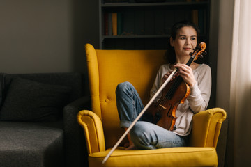 Pretty violinist woman sits with a violin in a soft chair at home looking out the window. Girl is posing with musical instrument. © dikushin