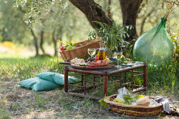 Romantic picnic under olive tree. Delicious italian meal served on a wooden table. Baskets with food, branches in glass jar. Sunny autumn day. Italy, Tuscany - obrazy, fototapety, plakaty