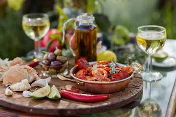 Romantic italian lunch outside for a couple: copper pan with delicious and spicy fried shrimps with herbs and garlic, wine, bread, olives. Bottle of olive oil. Luxury lifestyle, gourmet food