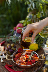 Girl is squeezing slice of lemon on delicious and spicy shrimps fried in copper pan with herbs, tomatoes and garlic. Wine, olives on the table. Luxury lifestyle, gourmet food for lunch