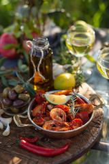 Romantic italian lunch outside for a couple: copper pan with delicious and spicy fried shrimps with herbs and garlic, wine, bread, olives. Bottle of olive oil. Luxury lifestyle, gourmet food