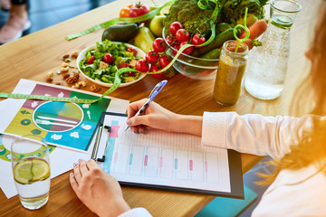 Woman dietitian in medical uniform with tape measure working on a diet plan sitting with different healthy food ingredients in the green office on background. Weight loss and right nutrition concept - obrazy, fototapety, plakaty