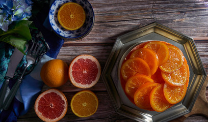 Top view and close-up of a cold homemade orange pie with candied orange on the surface and orange jam on a metal plate, accompanied by grapefruit, fresh oranges and flowers on a wooden background.