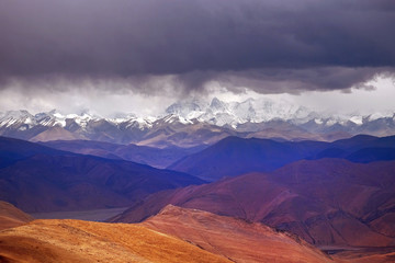 Panoramic view of the brown Himalaya Mountains, against the mountain range covered by snow and a dramatic sky.