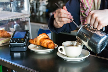 Barista female hands pouring hot milk on a cup to make a cappuccino with a croissant and a contactless post in the background.
