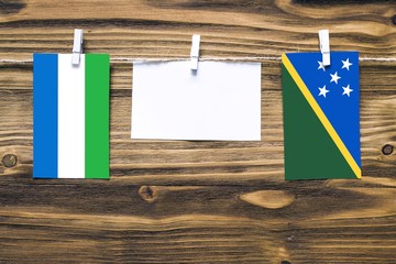 Hanging flags of Sierra Leone and Solomon Islands attached to rope with clothes pins with copy space on white note paper on wooden background.Diplomatic relations between countries.