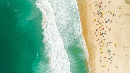 Fototapeta na wymiar Aerial locked shot of waves breaking on the shore. Colourful beach umbrellas and people enjoying the summer.